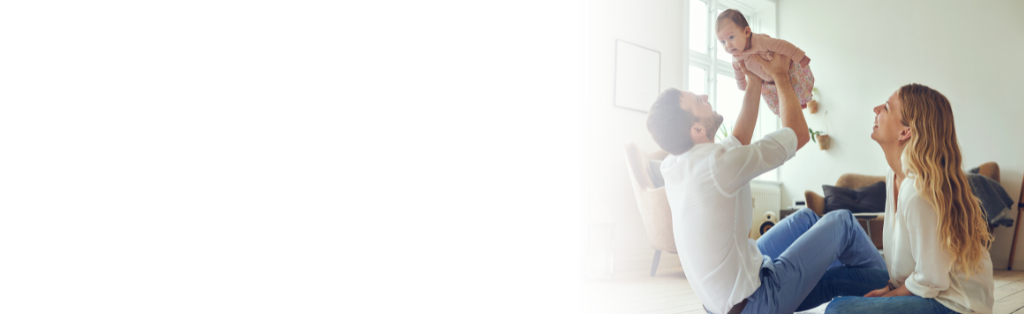 A mom and dad are sitting on the floor, the dad is playing with the baby and has her raised over him